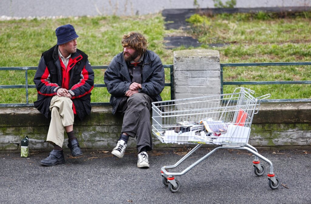 A homeless man sitting on a bench with a man trying to help him., with a grocery cart nearby.