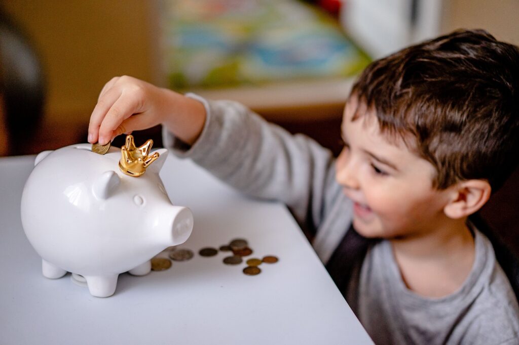 A smiling boy putting coins into a piggybank with a crown on it.