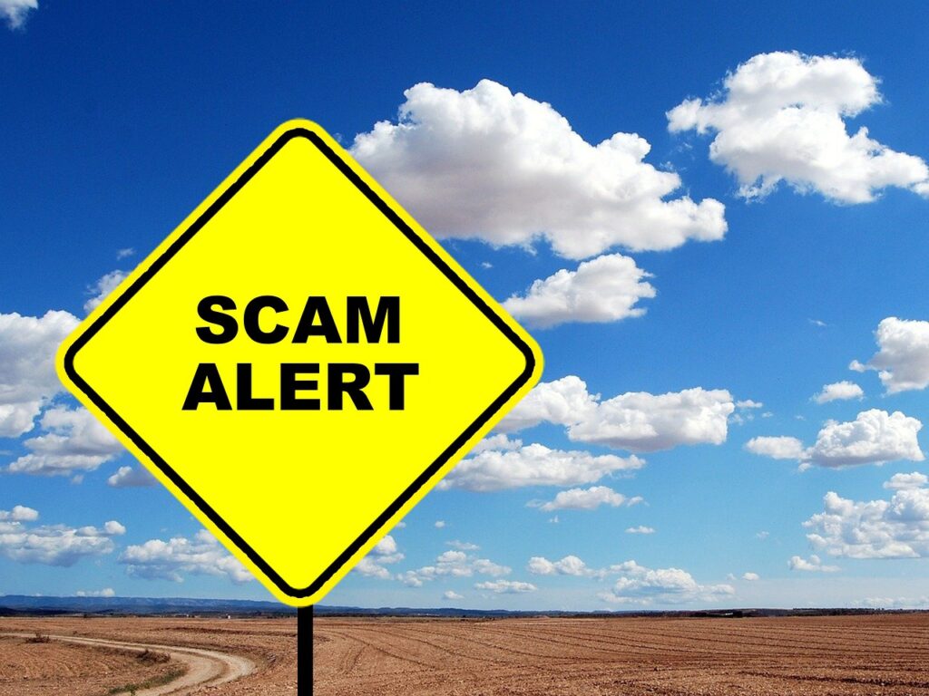 A yellow diamond shaped sign with black letters saying Scam Alert.