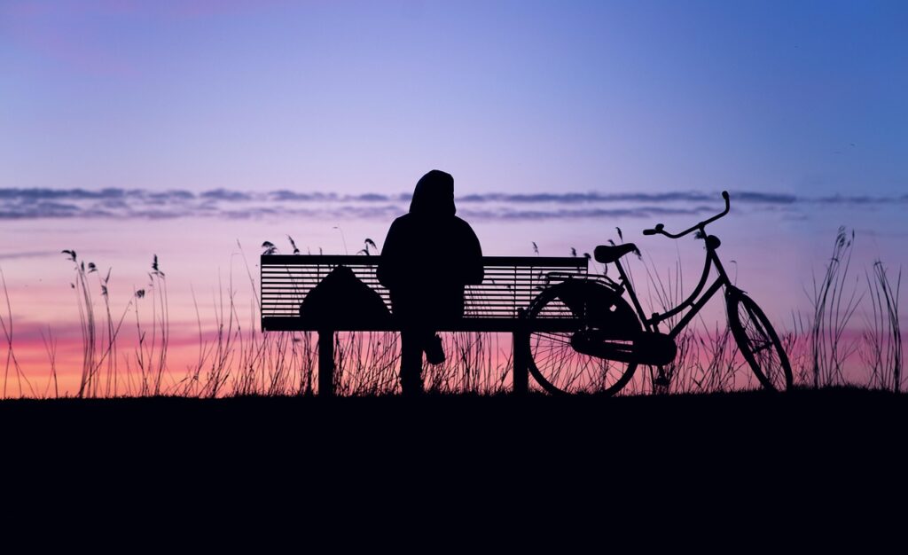 A person sitting on a bench with a bike nearby looking at a sunset.