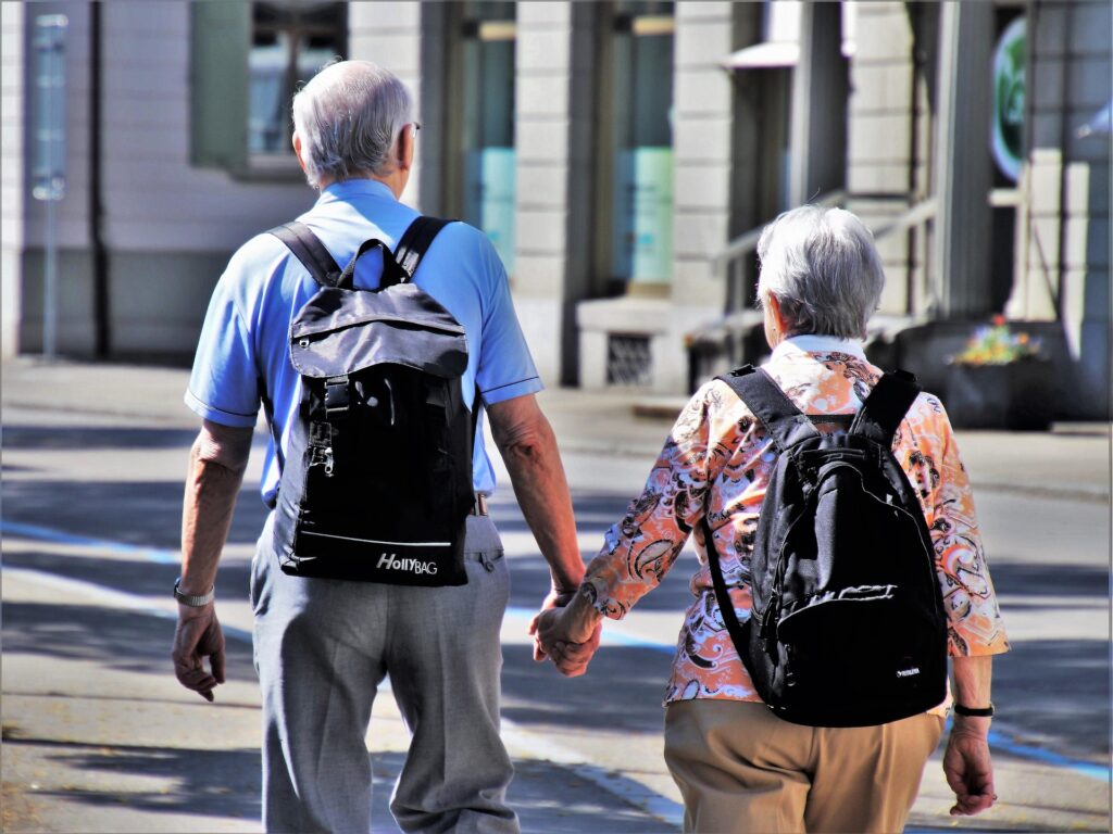 Grey headed man and a woman holding hands while walking in front of  a school with backpacks on.