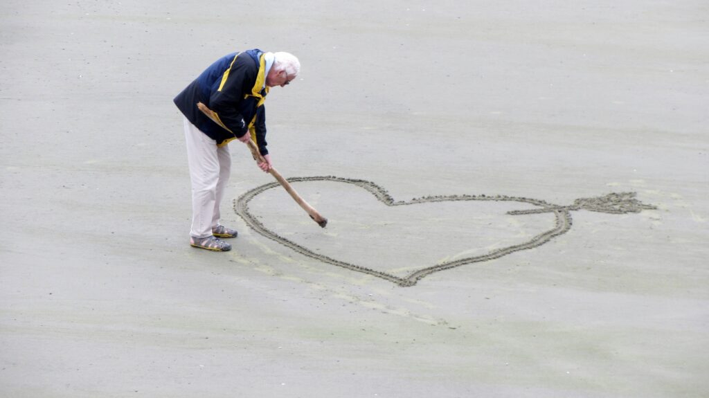 An old man drawing a heart with an arrow through it on the beach.